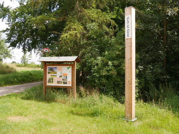 Bibel-Pfahl 20 - Borgentreich Ein hoher Holzpfahl mit der Aufschrift "Gott ist Geist" steht neben einer Infotafel am Wegesrand.
