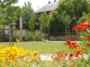 Bibel-Pfahl 21 - Manrode Ein gepflegter Gartenbereich mit bunten Blumen, jungen Bäumen und einem Bibel-Pfahl im Zentrum.