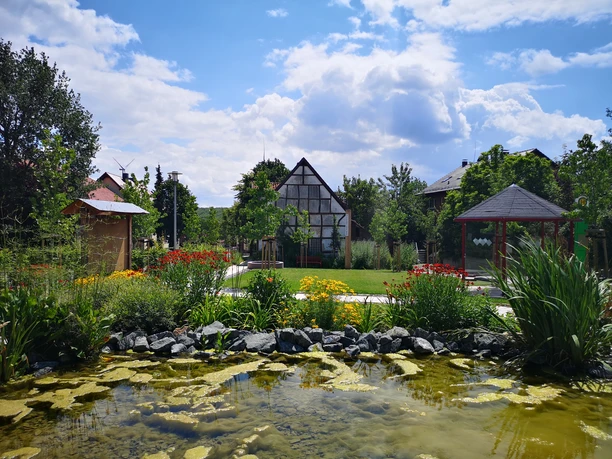 Fachwerkhaus mit üppigem Garten und Teich, umgeben von blühenden Pflanzen und vereinzelten Bäumen.