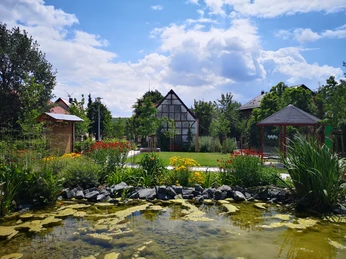 Bibel-Pfahl 21 - Manrode Fachwerkhaus mit üppigem Garten und Teich, umgeben von blühenden Pflanzen und vereinzelten Bäumen.