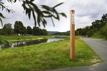 Bibel-Pfahl 28 - Corvey / Hafen Ein hölzerner Pfahl mit Bibelvers an einem Weg entlang eines Flusses, umgeben von grüner Landschaft.