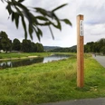 Bibel-Pfahl 28 - Corvey / Hafen Ein hölzerner Pfahl mit Bibelvers an einem Weg entlang eines Flusses, umgeben von grüner Landschaft.