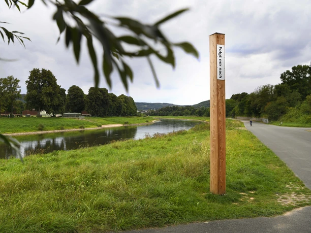 Bibel-Pfahl 28 - Corvey / Hafen Ein hölzerner Pfahl mit Bibelvers an einem Weg entlang eines Flusses, umgeben von grüner Landschaft.