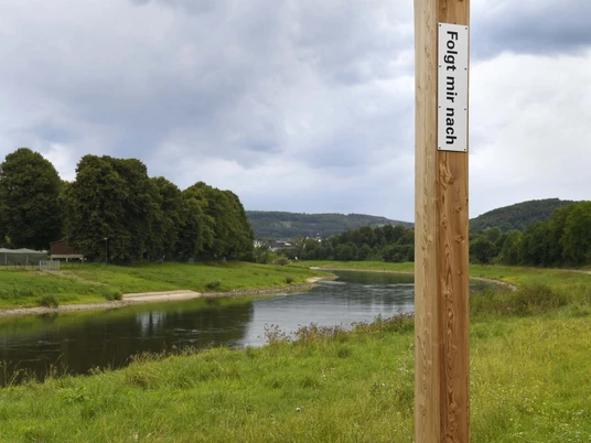 Holzpfahl mit Aufschrift "Folgt mir nach!" nahe einem Flussufer, bewölkter Himmel im Hintergrund.