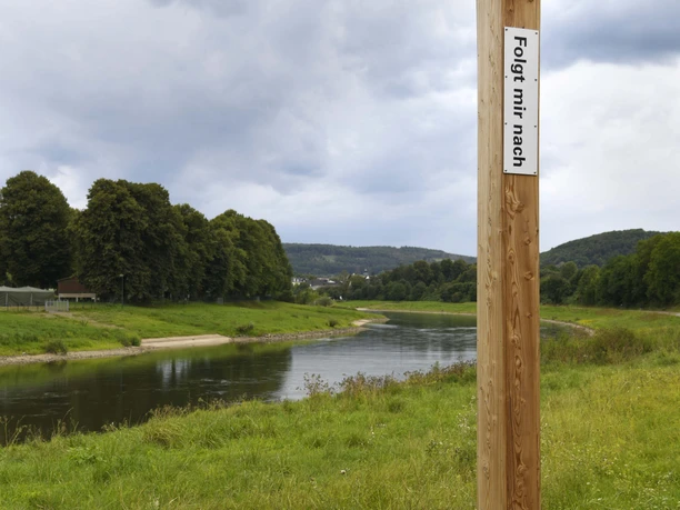 Holzpfahl mit Aufschrift "Folgt mir nach!" nahe einem Flussufer, bewölkter Himmel im Hintergrund.