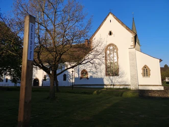 Ein weißes Kirchengebäude mit hohen Fenstern und spitzem Turm in Brenkhausen, umgeben von Bäumen.