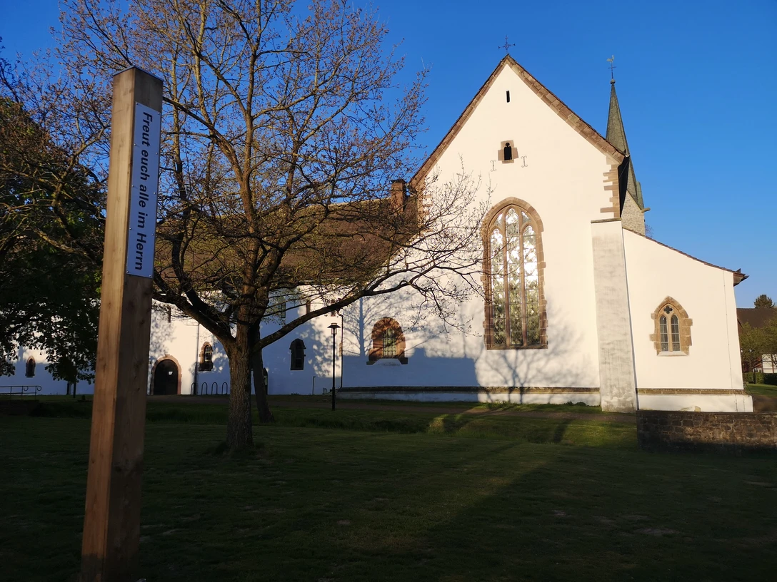 Bibel-Pfahl 30 - Brenkhausen Ein weißes Kirchengebäude mit hohen Fenstern und spitzem Turm in Brenkhausen, umgeben von Bäumen.