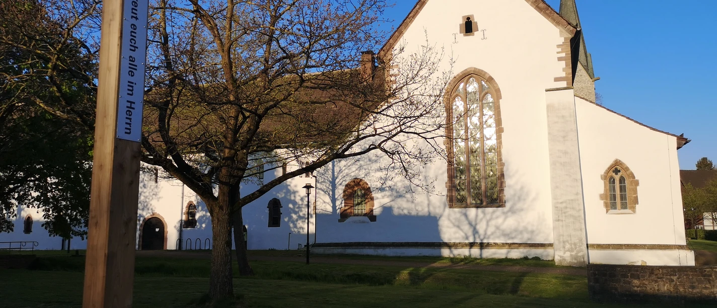 Bibel-Pfahl 30 - Brenkhausen Ein weißes Kirchengebäude mit hohen Fenstern und spitzem Turm in Brenkhausen, umgeben von Bäumen.