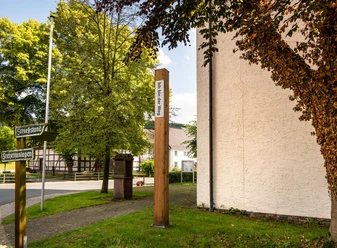 Bibel-Pfahl 31 nahe einer Kirche in Ovenhausen, von Bäumen und einem Wegweiser umgeben.
