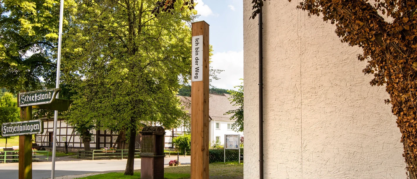 Bibel-Pfahl 31 nahe einer Kirche in Ovenhausen, von Bäumen und einem Wegweiser umgeben.