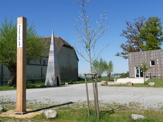 Ein Holzpfahl mit biblischem Text steht vor moderner Architektur und jungen Bäumen in Marienmünster.