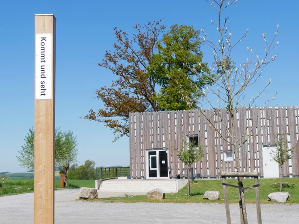 Ein hölzerner Pfahl mit der Aufschrift "kommt und seht" vor modernem Gebäude auf Schotterplatz.