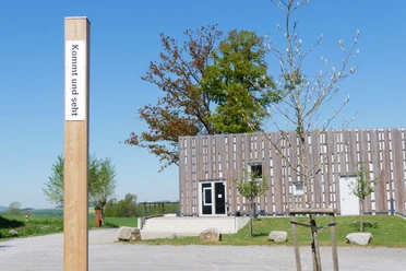 Bibel-Pfahl 33 - Abtei Marienmünster Ein hölzerner Pfahl mit der Aufschrift "kommt und seht" vor modernem Gebäude auf Schotterplatz.