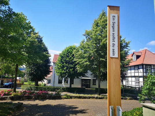 Ein hölzerner Pfahl mit der Aufschrift "Ein Segen sollst du sein" vor historischen Fachwerkhäusern.