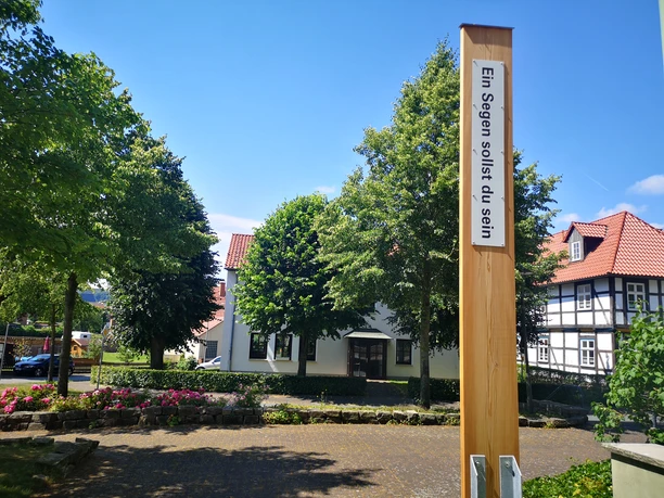 Ein hölzerner Pfahl mit der Aufschrift "Ein Segen sollst du sein" vor historischen Fachwerkhäusern.