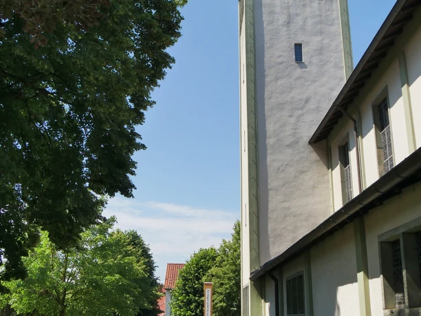 Hoher Kirchturm mit grüner Verkleidung neben einem schattigen Gehweg und Bäumen an einem sonnigen Tag.