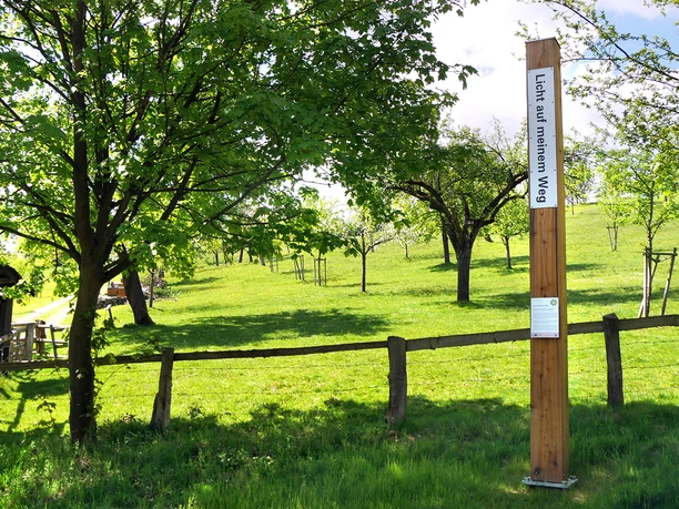 Ein hoher Pfahl mit Bibelverse in grüner Wiesenlandschaft, umgeben von Bäumen und einem Zaun.