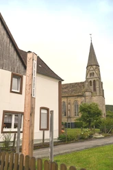 Bibel-Pfahl 37 - Nieheim Holzpfahl mit Bibelvers vor einem Wohnhaus, im Hintergrund eine Kirche mit hohem Turm.