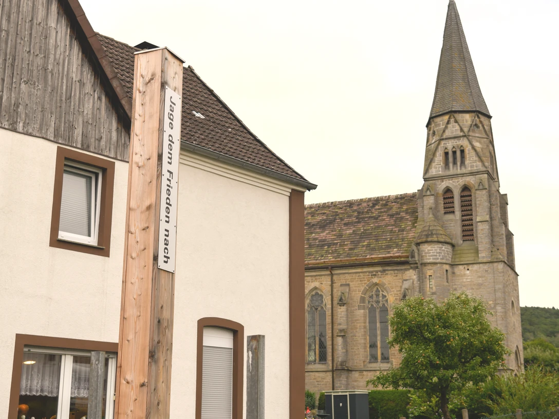 Bibel-Pfahl 37 - Nieheim Holzpfahl mit Bibelvers vor einem Wohnhaus, im Hintergrund eine Kirche mit hohem Turm.