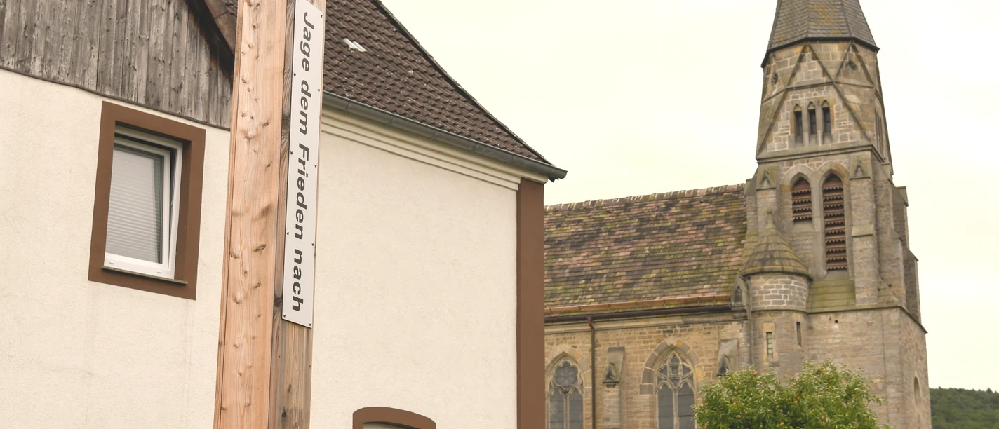 Bibel-Pfahl 37 - Nieheim Holzpfahl mit Bibelvers vor einem Wohnhaus, im Hintergrund eine Kirche mit hohem Turm.