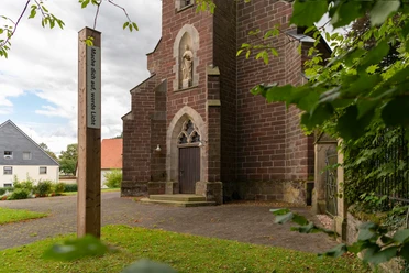 Bibel-Pfahl 40 - Merlsheim Ein Bibel-Pfahl mit der Inschrift "Möge dein Wort leuchten" steht vor einer alten Backsteinkirche.