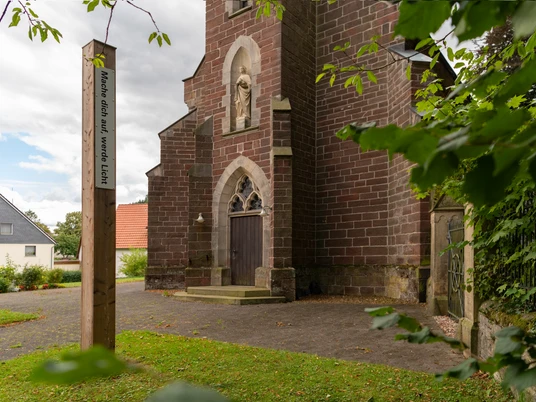 Bibel-Pfahl 40 - Merlsheim Ein Bibel-Pfahl mit der Inschrift "Möge dein Wort leuchten" steht vor einer alten Backsteinkirche.