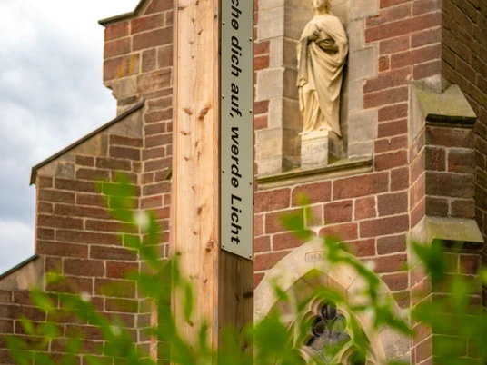 Bibel-Pfahl 40 - Merlsheim Ein Holzpfahl mit der Inschrift "Mache dich auf, werde Licht" steht vor einer Kirchenfassade.