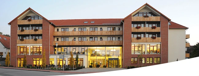 landhotel.jpg Ein mehrstöckiges, modernes Landhotel mit rotgedeckten Giebeln und Balkonen, umgeben von Bäumen.