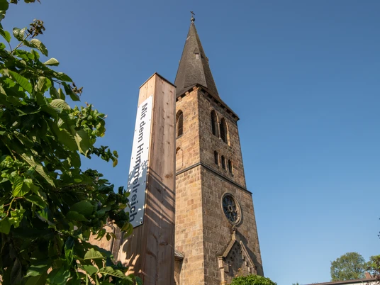 Bibel-Pfahl 41 - Bad Driburg Historische Kirche mit hohem Turm, flankiert von einem modernen Pfahl mit Bibelzitat, klarer Himmel.