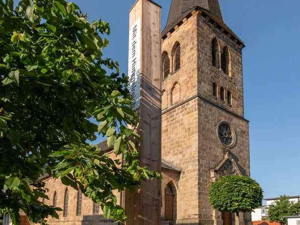 Bibel-Pfahl 41 - Bad Driburg Pfahl mit Bibelvers vor historischer Kirche, umgeben von grünem Rasen und blauem Himmel.