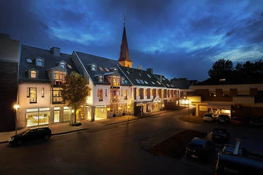 Hotelgebäude bei Dämmerung, beleuchtet, mit Kirchturm im Hintergrund und geparkten Autos vorne.