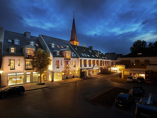 Hotelgebäude bei Dämmerung, beleuchtet, mit Kirchturm im Hintergrund und geparkten Autos vorne.