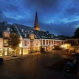 Reuter_1.jpg Hotelgebäude bei Dämmerung, beleuchtet, mit Kirchturm im Hintergrund und geparkten Autos vorne.