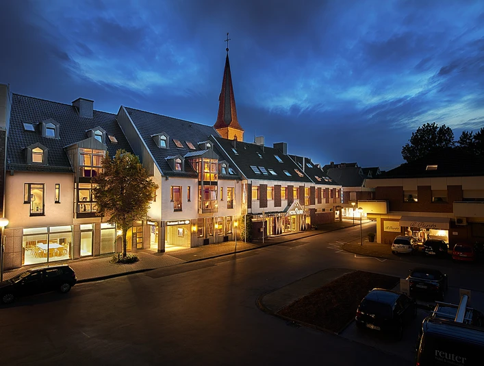 Reuter_1.jpg Hotelgebäude bei Dämmerung, beleuchtet, mit Kirchturm im Hintergrund und geparkten Autos vorne.