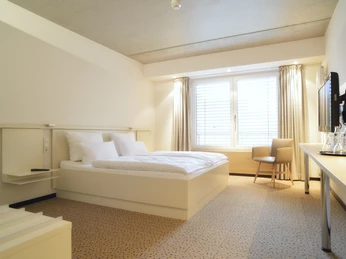 Reuter_3 Helles, modernes Hotelzimmer mit Doppelbett, Sessel, Schreibtisch und großem Fenster mit Tageslicht.