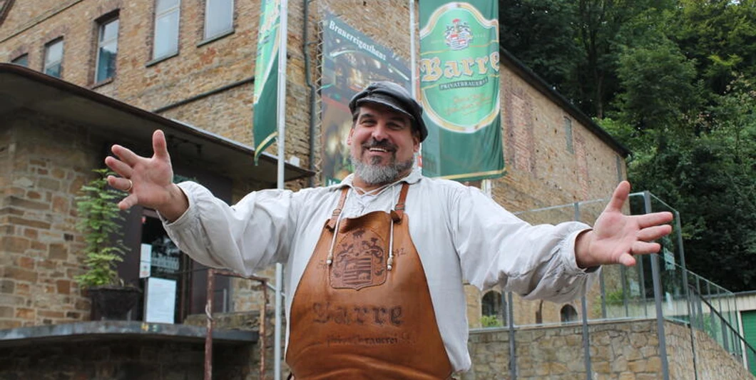 Barres Brauwelt Ein lachender Mann in Leder-Schürze steht vor einem Gebäude der Brauerei Barre in Löhne.