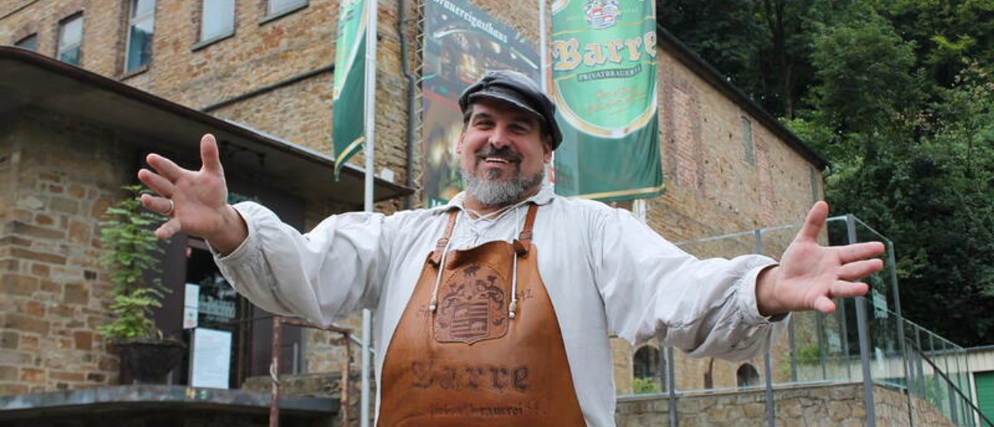 Barres Brauwelt Ein lachender Mann in Leder-Schürze steht vor einem Gebäude der Brauerei Barre in Löhne.