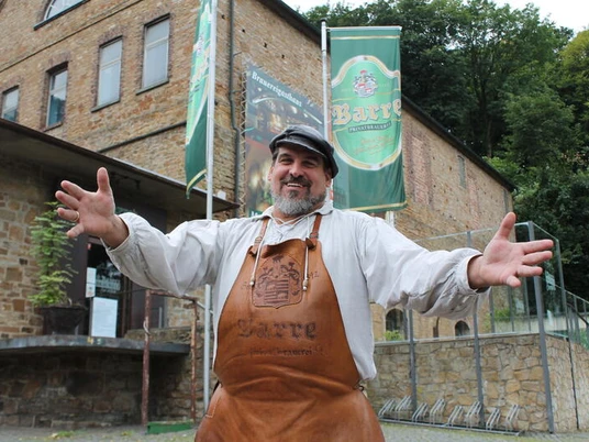 Barres Brauwelt Ein lachender Mann in Leder-Schürze steht vor einem Gebäude der Brauerei Barre in Löhne.