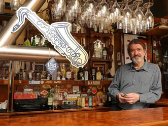 Blue-Mojo Mann steht in einer Bar, umgeben von Gläsern und Flaschen. Im Hintergrund eine Leuchtreklame.