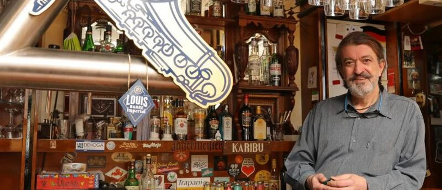 Blue-Mojo Mann steht in einer Bar, umgeben von Gläsern und Flaschen. Im Hintergrund eine Leuchtreklame.