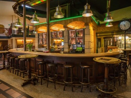 Brauhaus Espelkamp Große, rustikale Bar mit Holzhockern, umgeben von warmem Licht. Industrielle Lampen hängen über der Theke.
