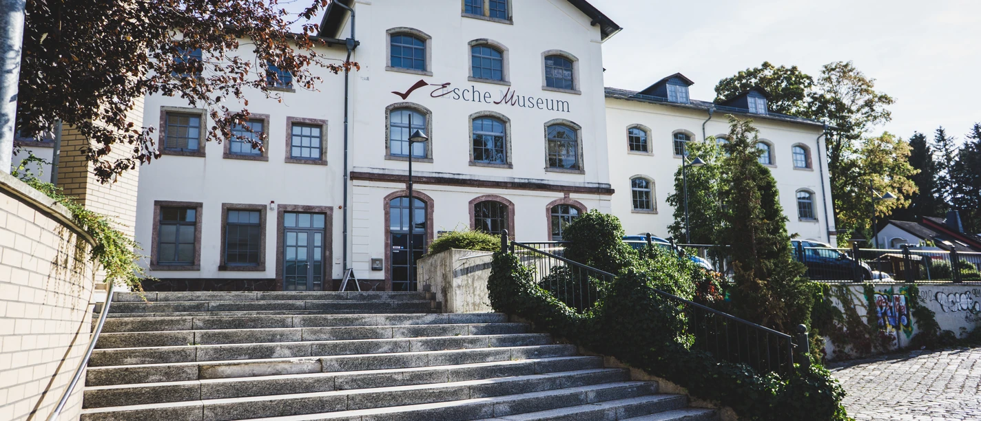 Außengebäude Esche-Museum.jpg Weißes mehrstöckiges Gebäude mit Schildern, Treppen und Pflanzen im Vordergrund.
