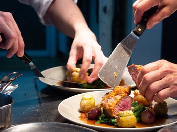 Reuter_8 Köche garnieren sorgfältig gegrilltes Fleisch mit Gemüse auf stilvoll arrangierten Tellern in einer Küche.