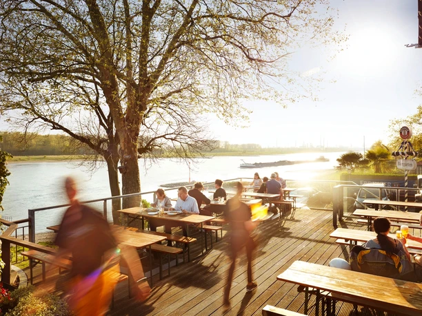 Haus Rheinblick Ott Eine belebte Terrasse am Flussufer des Rheins mit Menschen, die an Tischen speisen und genießen.