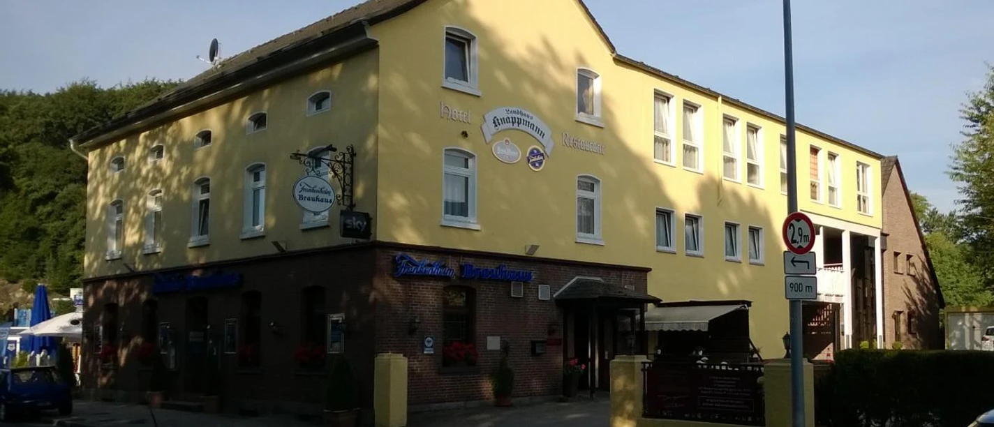 Gelbes Gebäude mit Gasthaus und Hotel in sonniger Umgebung, umgeben von Bäumen und Straße.