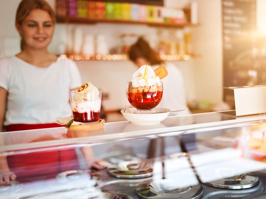 Eiscafé art & wEISe Zwei extravagante Eisbecher mit Toppings auf einer Theke, bedient von einer lächelnden Person im Eiscafé.