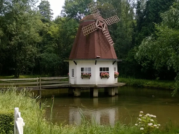 Restaurant Heidberger Mühle Eine historische Mühle mit rotem Dach und Flügeln steht idyllisch von Natur umgeben an einem Teichrand.