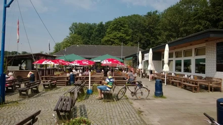 Se(h)-Restaurant Unterbacher See Sonnige Terrasse eines Restaurants am See mit rot-weißen Sonnenschirmen, Holzsitzbänken und Fahrrädern.
