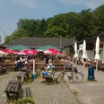 Se(h)-Restaurant Unterbacher See Sonnige Terrasse eines Restaurants am See mit rot-weißen Sonnenschirmen, Holzsitzbänken und Fahrrädern.