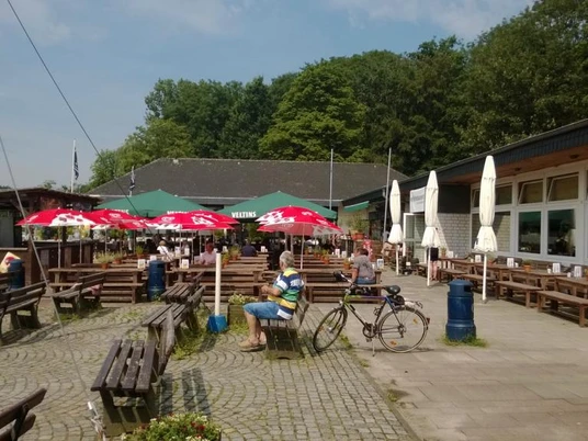 Se(h)-Restaurant Unterbacher See Sonnige Terrasse eines Restaurants am See mit rot-weißen Sonnenschirmen, Holzsitzbänken und Fahrrädern.